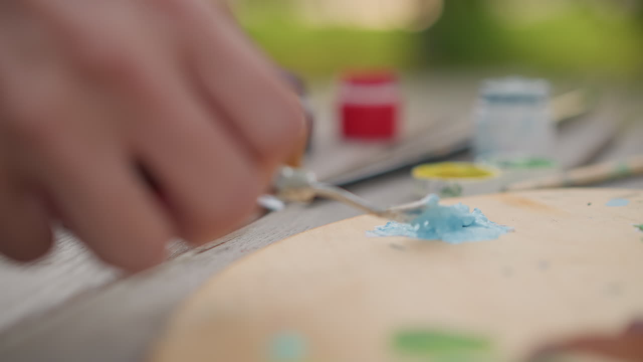 屋外のパレットで青い顔料を混ぜ、アーティストが金属製のナイフを使って絵の具の瓶やチューブを削ったり盛ったりしている様子。背景はソフトなボケ味でぼやけ、自然な夏の光が差し込み、穏やかで瞑想的な雰囲気です。