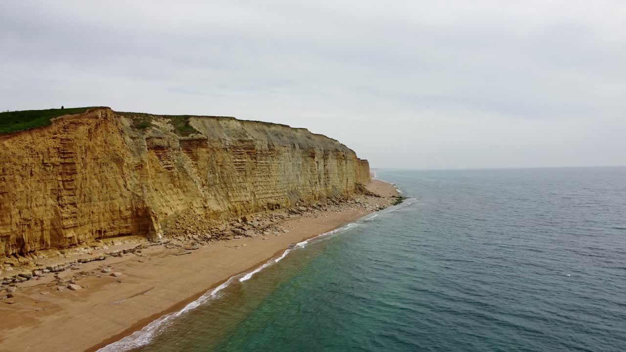 Aerial footage rotating around a sheer cliff face on a Jurassic Coast Beach in Dorset. The footages starts moving quickly to reveal the full size and scope of this amazing cliff on a cloudy day.