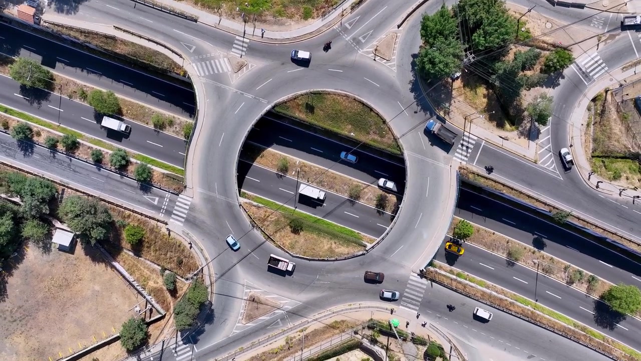 Ascending spiral aerial shot directly above urban roundabout with cars and trucks flowing around circular ring road intersection revealing the perfect geometric traffic pattern