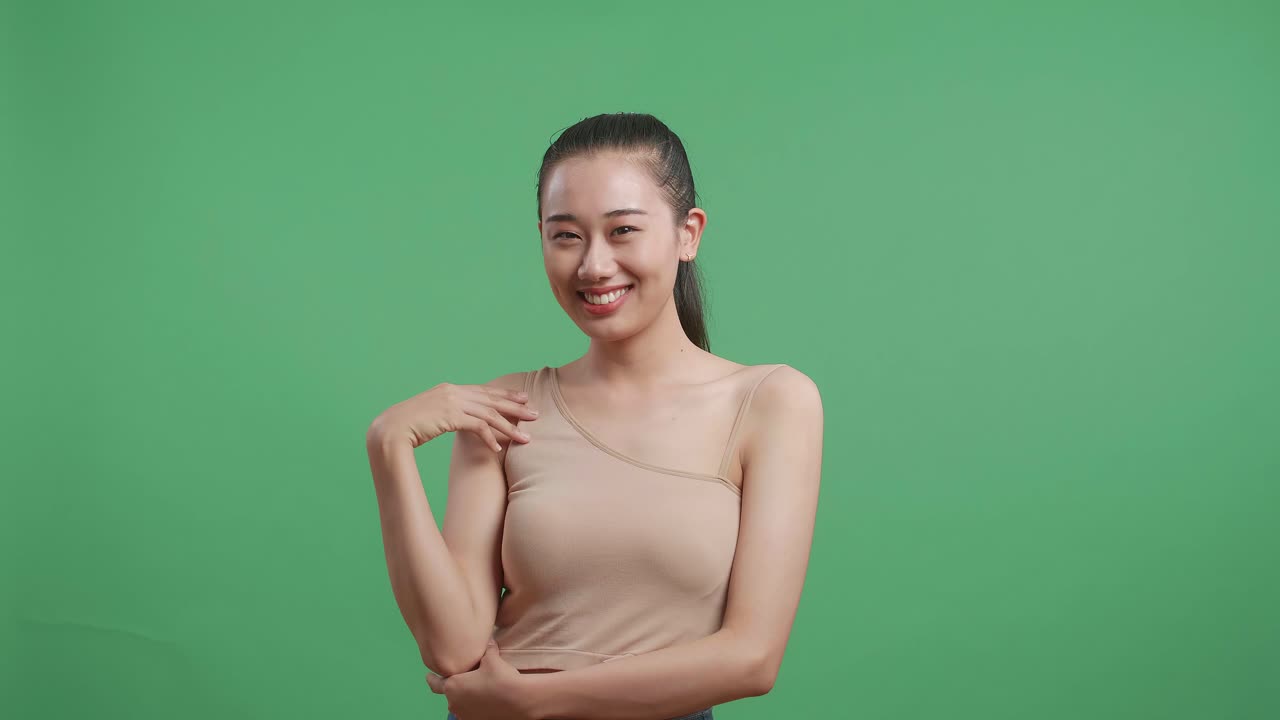 아름다운 아시아 여성은 그녀의 피부를 만지고 스튜디오에서 녹색 화면 배경에 카메라를 보고 미소 짓고 있습니다.