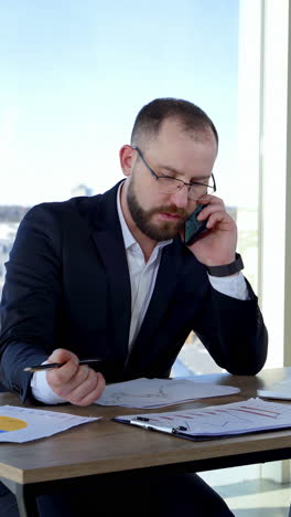 business worker at his workplace. confident man in glasses sitting at the desk with a laptop and talking on a phone looking into papers in office. vertical video
