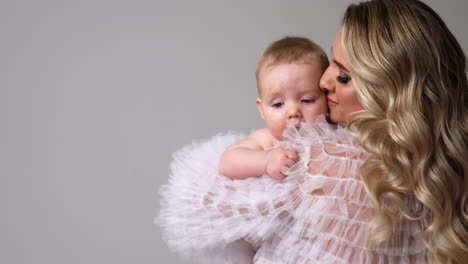 beautiful loving mother kissing her adorable little baby. woman embracing her child and turning around with him. white backdrop.