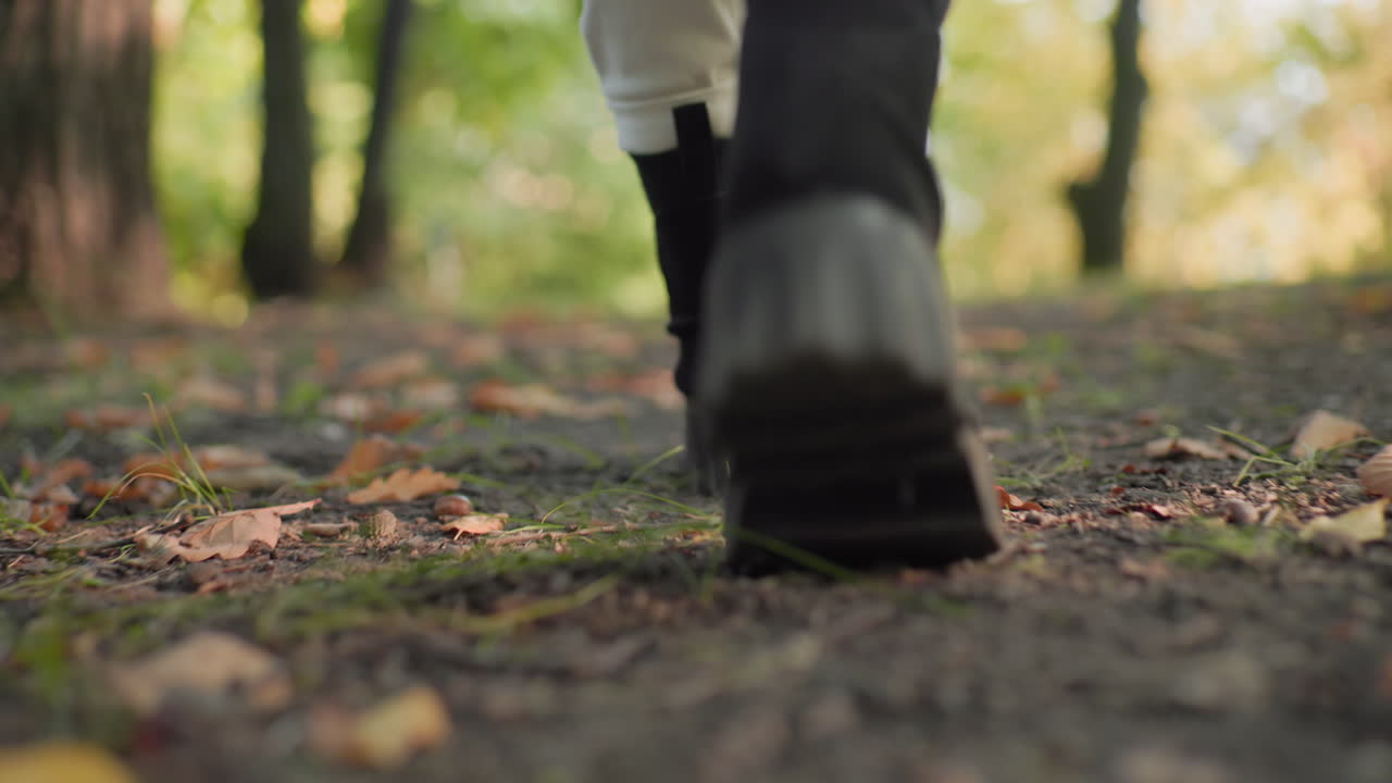 Back leg view of forest visitor walking in sturdy boots, slow relaxed steps along earthy path, heel rising from dirt, crunchy leaves and acorns underfoot, soft bokeh woods and autumn light