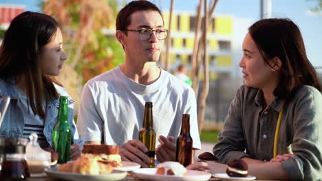 group of three japanese friends talking and drinking beer while sitting at table outdoors in a sunny day 2