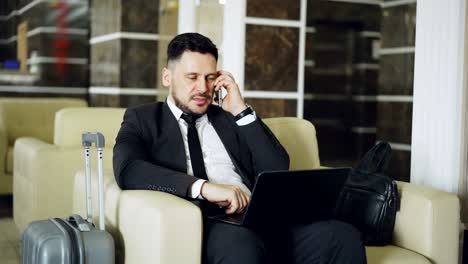 tilt up of confident businessman talking mobile phone and using laptop computer while sits on armchair in luxury hotel with luggage near him. travel, business and people concept