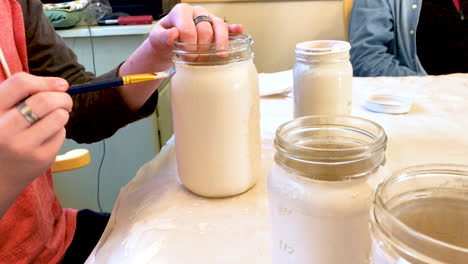 Closeup-of-person-sitting-at-a-table-in-a-home-and-painting-mason-jars-for-decoration-at-an-upcoming-social-event