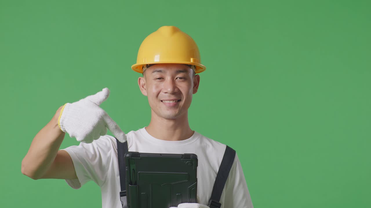 Close Up Of Asian Man Painter Wearing Safety Helmet Smiling And Pointing To A Tablet In His Hand While Standing In The Green Screen Background Studio