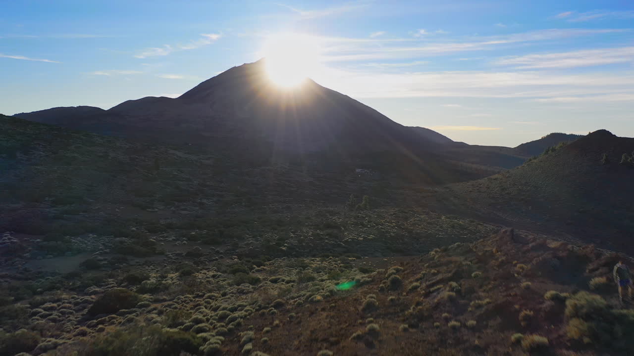 The Sun setting behing the summit of the Pico de Teide mountain on Canary Islands shining on the rocky hillside below with stones and bushes and a blue sky above. The Sun setting behing a mountain 4K.