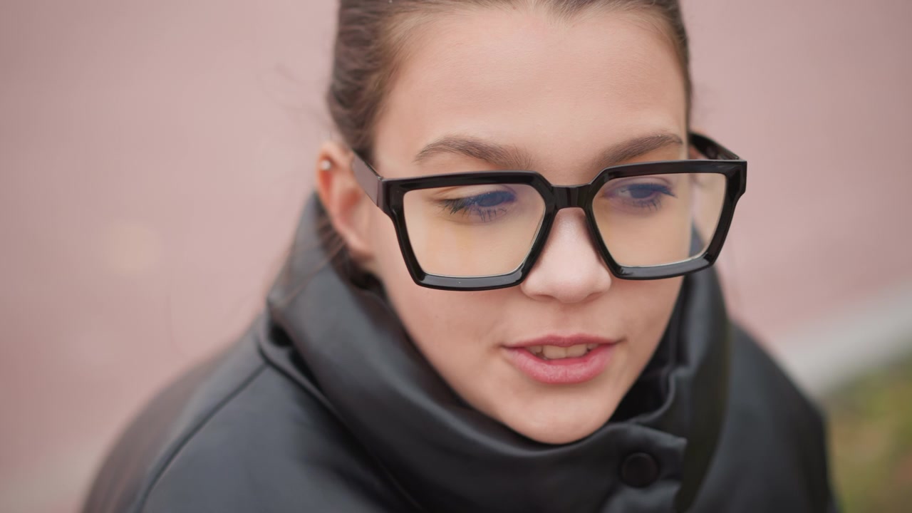 Primo piano di una giovane donna che indossa occhiali neri rettangolari e abiti scuri con i capelli raccolti all'indietro. La scena mantiene un'illuminazione soffusa per tutta la durata, concentrandosi sul viso del soggetto contro uno sfondo sfocato