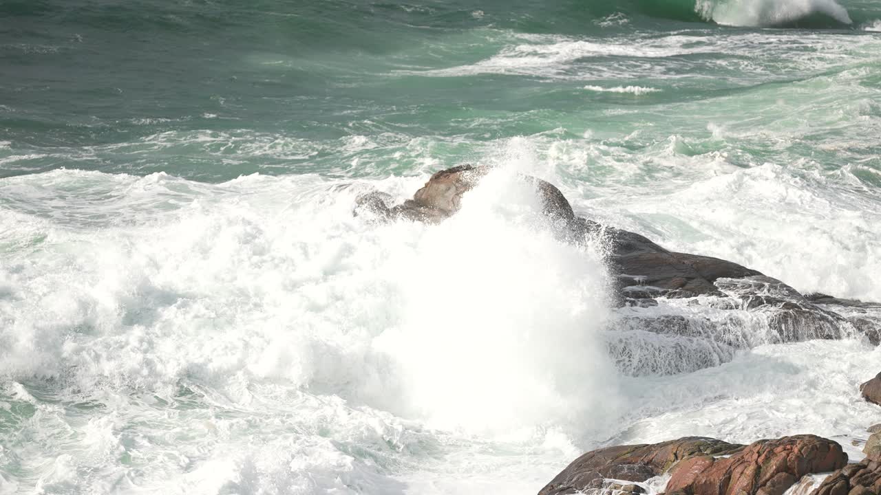Strong waves crash against large rocks in a coastal area, creating foam as they splash. The scene captures the beauty of nature during a serene sunset time