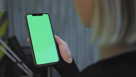 over shoulder shot of caucasian girl with blond hair sitting on sofa and holding smartphone with green screen in both hands at home.