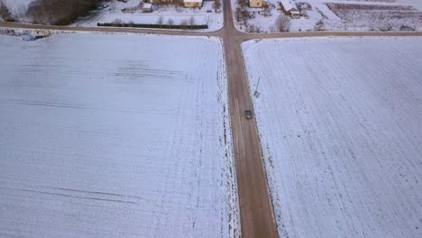 Birds-eye-view-of-vehicle,-moving-towards-rural-village-on-countryside-road,-amidst-snow-covered-fields