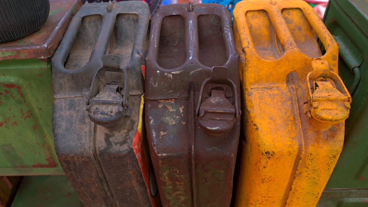 Un'inquadratura statica ravvicinata di tre taniche metalliche in stile militare usurate e arrugginite nei colori nero, marrone e giallo, che mostrano una texture invecchiata e dettagli metallici