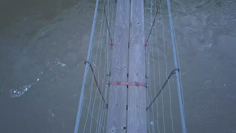 sketchy pov crossing narrow suspension foot bridge over swirling river