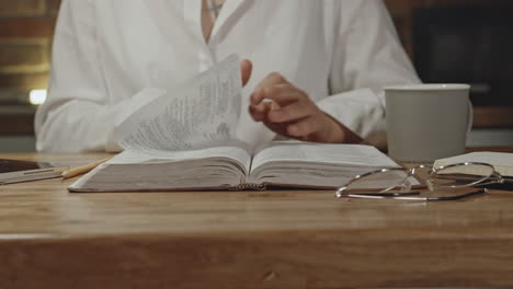 Woman-is-reading-and-flipping-the-pages-of-the-bible,-sitting-up-to-a-desk-with-glasses-and-coffee-cup