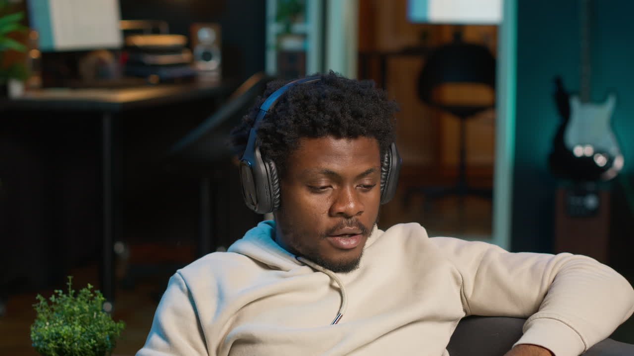 Black young adult attending a webinar on laptop from his living room, relaxing on his couch and listening on headset. Cheerful man multitasking in order to finish his daily tasks. Camera A.