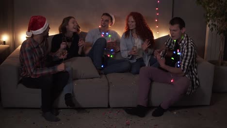 Cheerful-close-friends-celebrating,-having-time-together-at-home.-Happy-friends-sit-around-on-sofa-and-listen-to-guy-singing-and-playing-guitar.-Get-together-at-Christmas-time.-Muffled-light,-garland-lights