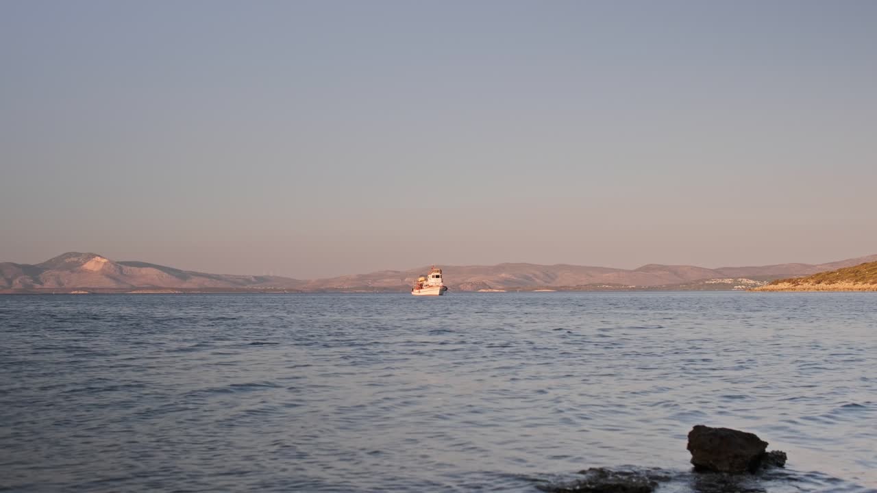 Cinematic footage of a pristine Blue Flag bay in Çeşme, Turkey. Swimmers and paddleboarders enjoy the clear, calm turquoise water with a lush green forest landscape in the background on a sunny day