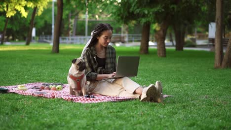 Junges-Mädchen,-Das-Mit-Ihrem-Laptop-Im-Freien-Auf-Dem-Rasen-Sitzt,-Und-Ein-Süßer-Kleiner-Mops-Sitzt-In-Der-Nähe