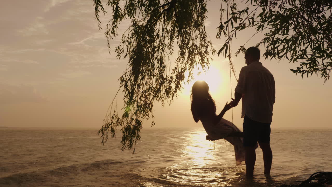 한 남자가 바다를 배경으로 그네를 타고 아내를 굴리고 있는 젊은 부부의 실루엣