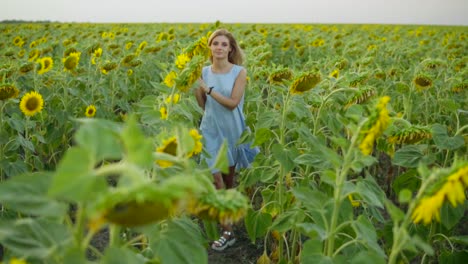 Portrait-of-a-young-woman-in-a-blue-dress-walking-in-the-sunflower-field,-looking-in-the-camera.-Beautiful-lady-enjoying-nature