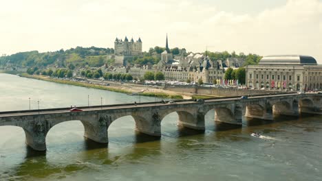 Aerial-shot-crossing-a-river-with-an-old-bridge-and-beautiful-chateau-in-the-background-in-France