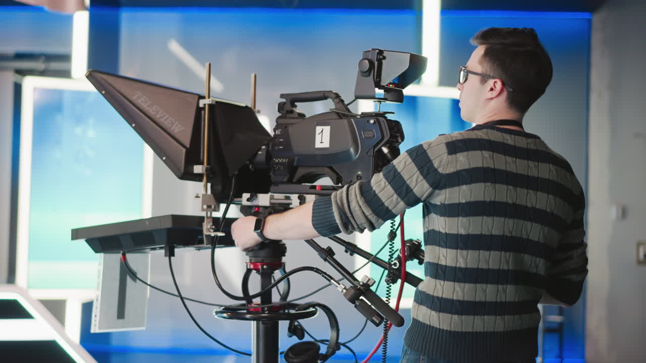 Man operating professional camera rig with teleprompter in broadcast studio. Camera setup with monitor and various controls. Operator wearing striped sweater, focused on filming process