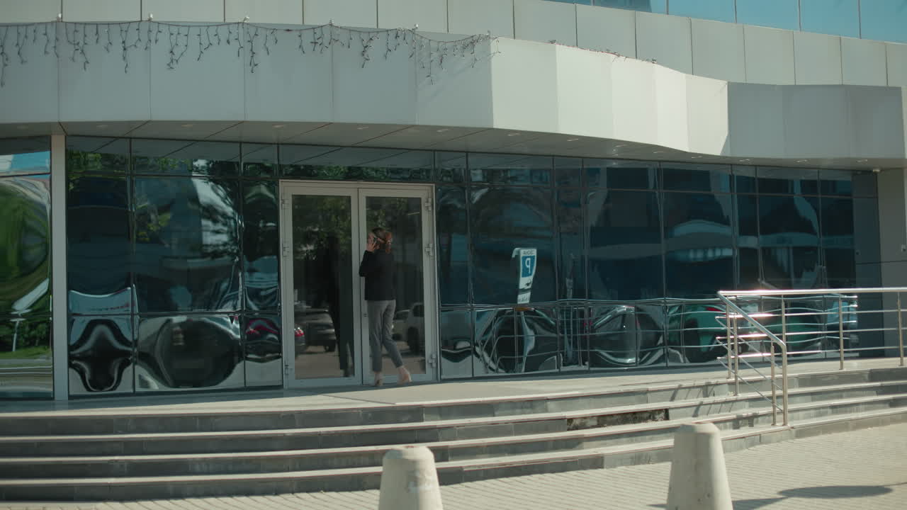 Woman holding handbag walking to work on phone call, entering modern building with glass facade reflecting parked cars and iron railing, showcasing professional lifestyle in urban environment