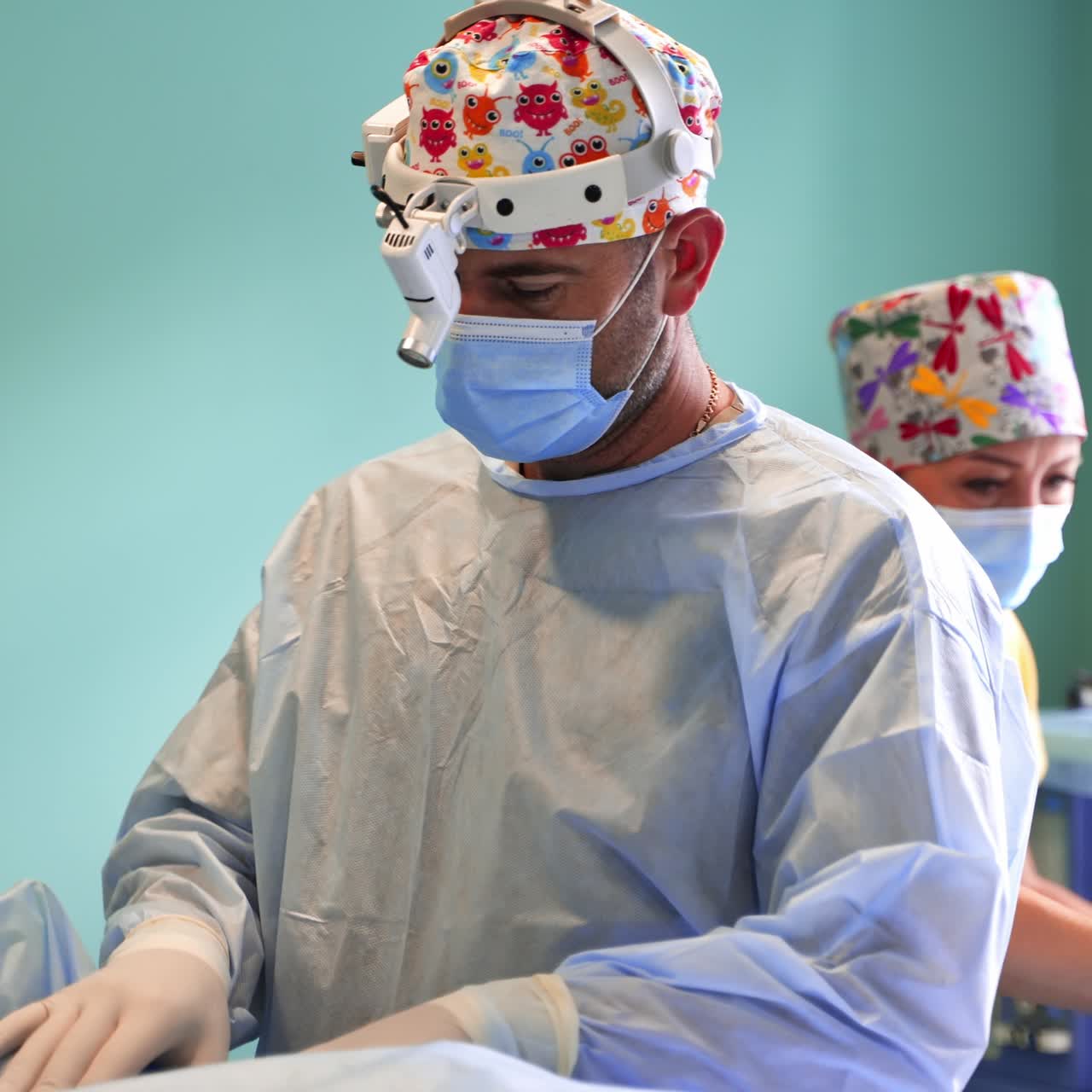 Adult male surgeon in funny bright hat and special device comes to a patient. Female assistant at backdrop
