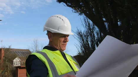un arquitecto senior examinando los planes de un gran edificio en un sitio de construcción en una calle residencial con tráfico en la carretera en el fondo