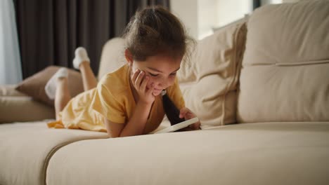 a-little-brunette-girl-lies-on-her-stomach-and-looks-at-the-screen-of-a-white-phone-on-a-light-brown-sofa-in-a-modern-apartment.-Little-girl-playing-a-game-or-watching-a-video-on-her-phone