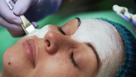 applying white face mask on female face. woman in blue protective cap having facial care at beautician's. extreme close up.