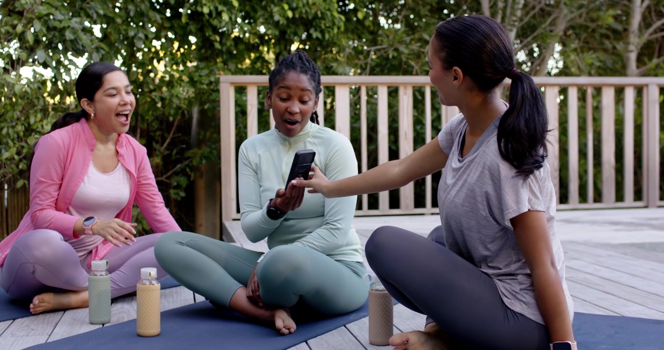 Diverse women on mats by pool taking selfie, right woman checking phone to share, center smiling