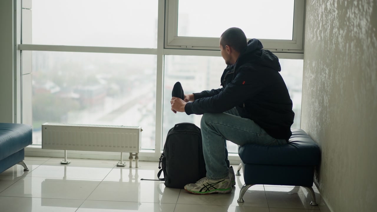 Man sits on bench by large windows putting on his shoe while looking out at city view. Man wears casual clothing and has luggage beside him in indoor waiting area with modern furnishings