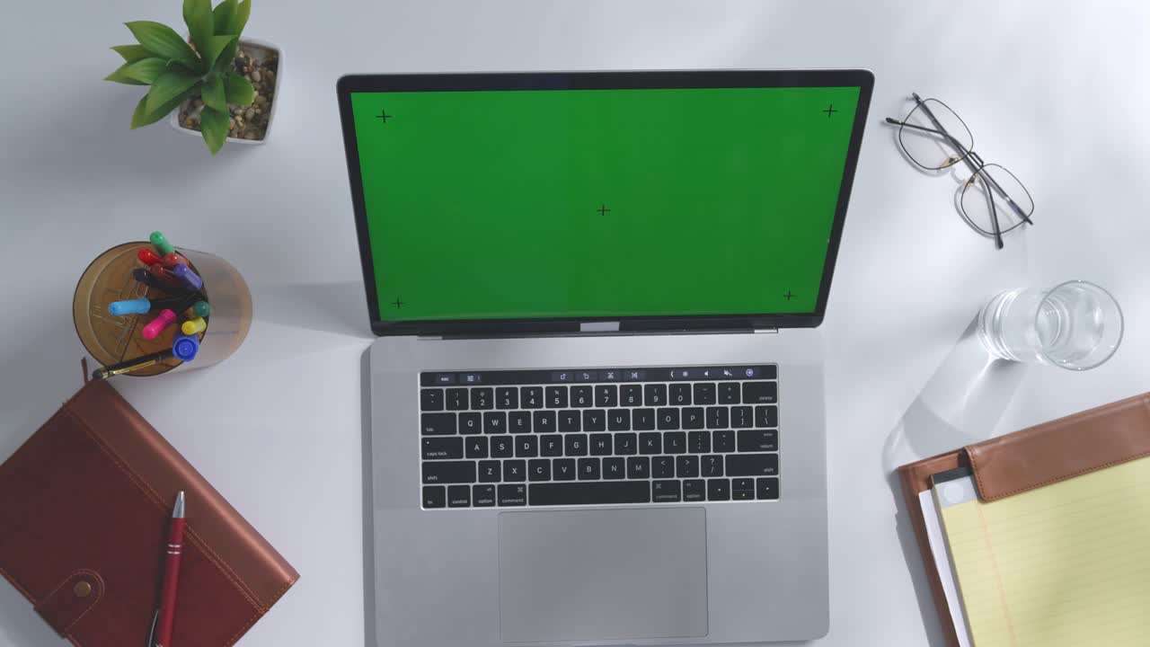 vista de arriba hacia abajo de una computadora portátil con pantalla verde simulada pantalla chromakey en un escritorio de oficina de madera al lado de la libreta con bolígrafos, gafas y un vaso de agua.