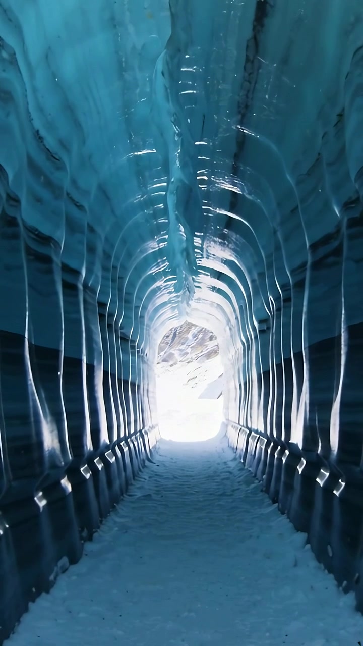 Video verticale: Telecamera in movimento lungo un tunnel di ghiaccio blu, luce del giorno che illumina stalattiti di ghiaccio e un sentiero innevato verso l'uscita