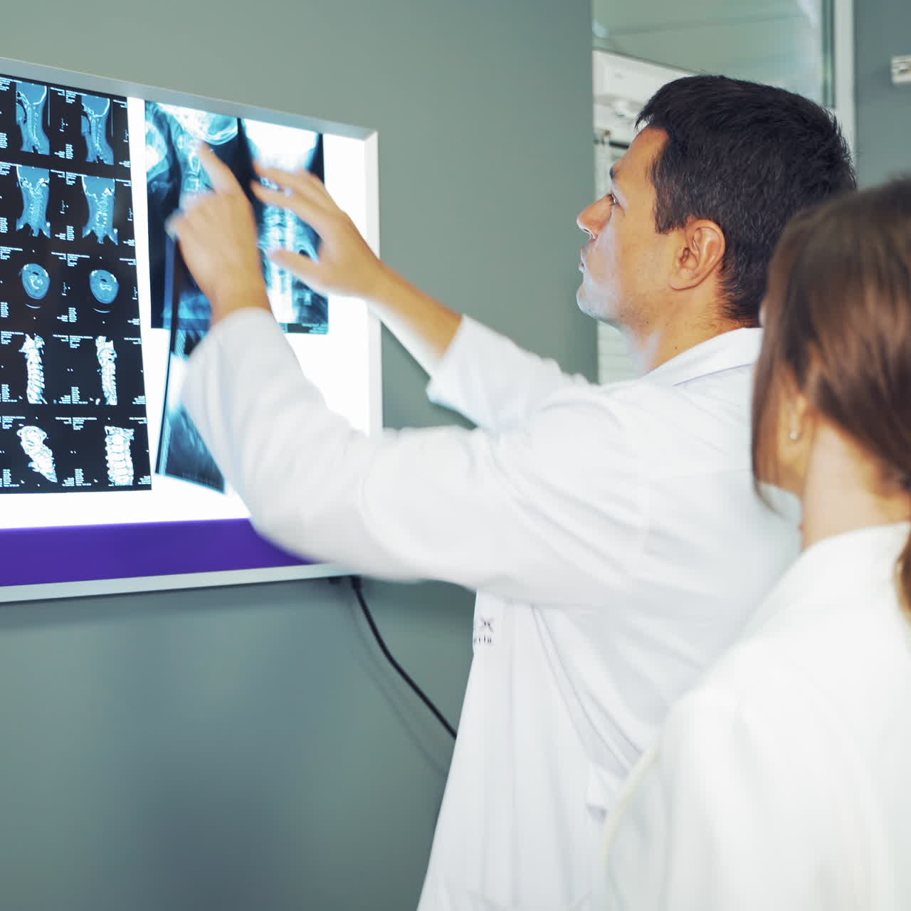 Male doctor is hanging a x-ray photograph on white board. Two medical coworkers in white uniform working with spinal tomography indoors in clinic.