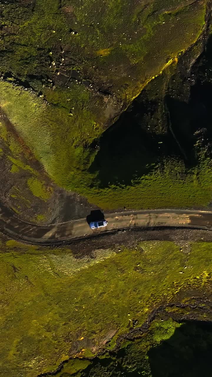 Ripresa aerea verticale dall'alto verso il basso di un SUV 4x4 su una pista sterrata accanto a un lago vulcanico scuro in Islanda, circondato da muschio verde intenso e sabbia nera