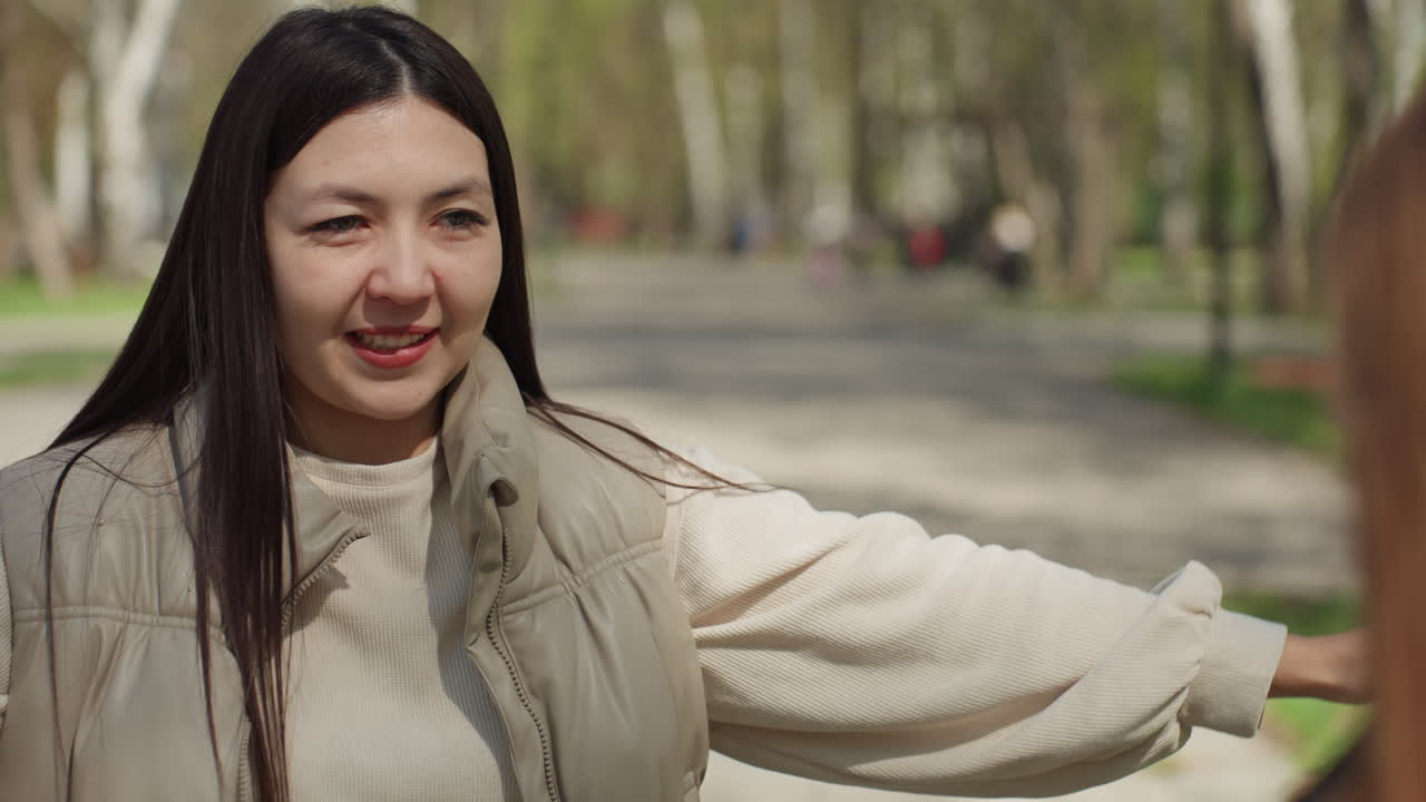 따뜻한 야외 인사, 아시아 여성이 친구를 따뜻하게 맞이함, 밝은 야외에서 두 여성이 즐겁게 재회함, 햇살 가득한 봄날에 아시아인과 백인 친구가 편안하게 재회함