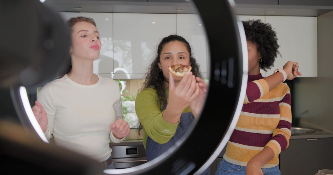 Diverse women filming pastries in kitchen, woman in white tasting, reacting under ring light camera