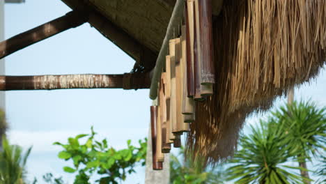 row of bamboo wind chimes hang on wooden poles from thatched roof of outdoor gazebo