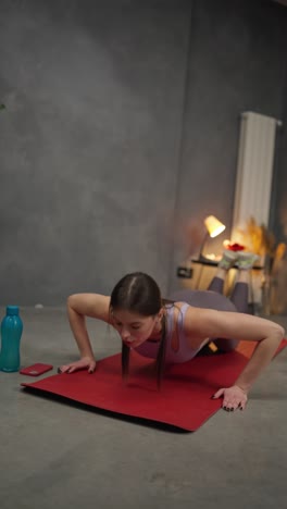 Vertical-video-Confident-brunette-girl-in-sports-summer-uniform-doing-push-ups-on-a-red-sports-mat-near-a-sports-bottle-of-water-in-a-modern-apartment-at-home