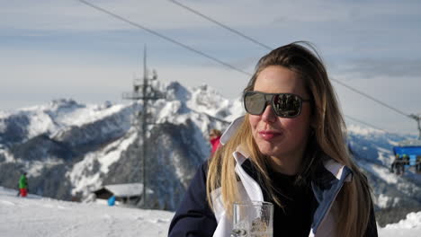 medium closeup of a caucasian woman with brown hair wearing sunglasses and drinking beer at a skiing resort with skiing cable cars and snow covered mountains in the background