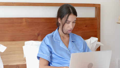 woman in blue pajamas using laptop in bed, looking thoughtfully out window, at home