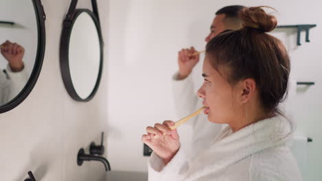 young family brushes teeth in bathroom. couple in terry bathrobes carry out hygienic routine standing before mirrors together at home. care routine