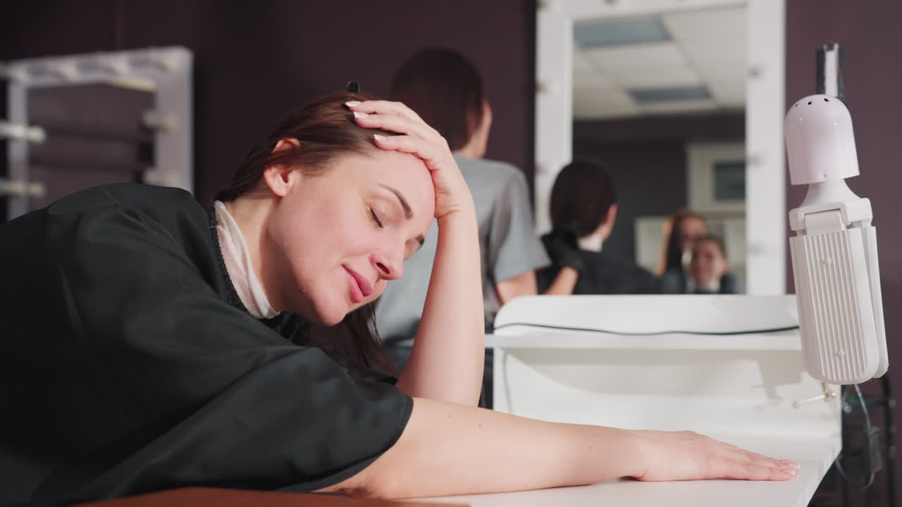 차분한 분위기의 미용실 데스크에서 일하는 직원, 은은한 조명 아래 조용히 앉아 있는 지친 접수 담당자, 평온한 미용실 환경과 부드러운 조명 속에서 손에 기댄 채 지쳐있는 프런트 데스크 직원