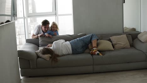 en la sala de estar cansada madre acostada en el sofá con el teléfono inteligente, adorable hija y padre de repente aparecen desde el sofá de atrás y