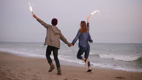 vista retrospectiva de una joven pareja multiétnica corriendo por el mar y sosteniendo velas brillantes durante la puesta del sol. disparo en cámara lenta