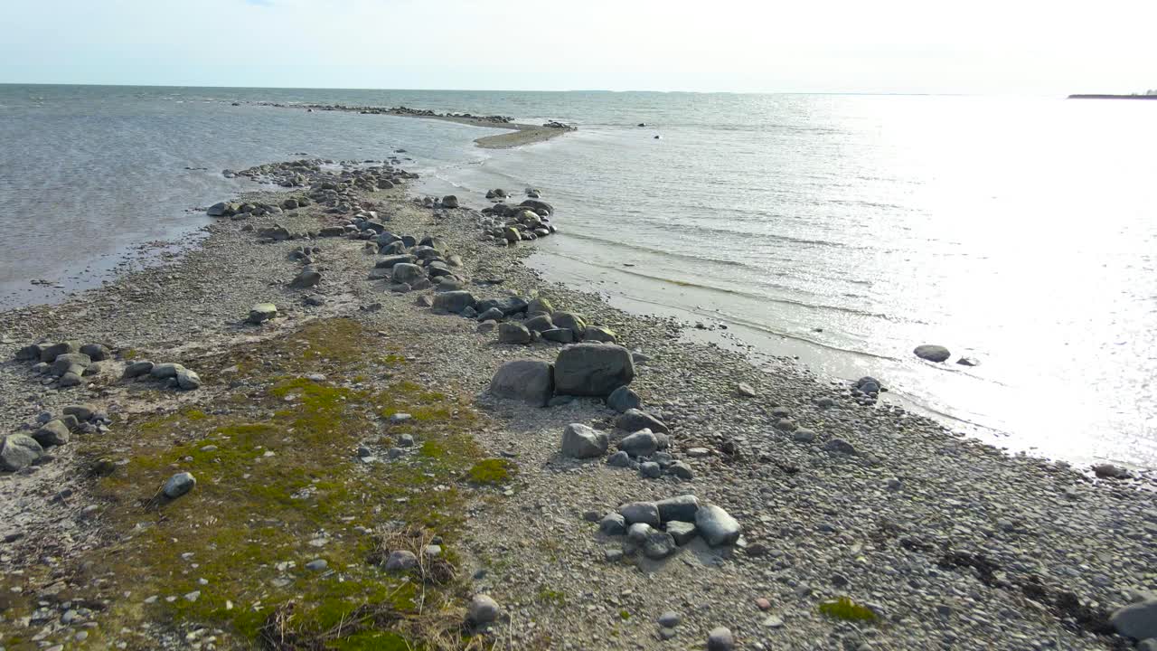 Flying low over illuminated coastal water and rocky shore while birds are flying by at Puhtulaiu peninsula, Estonia’s coastline. Rugged shoreline full of pebbles, sunlight reflection over sea surface
