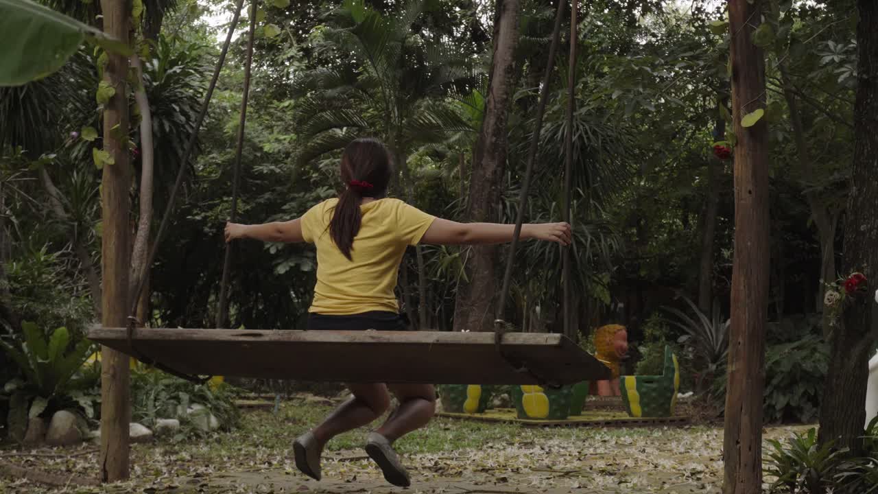 la espalda de una joven asiática en un columpio al aire libre, con los brazos extendidos preparándose mientras se mece de un lado a otro en un jardín en kanchanaburi, tailandia